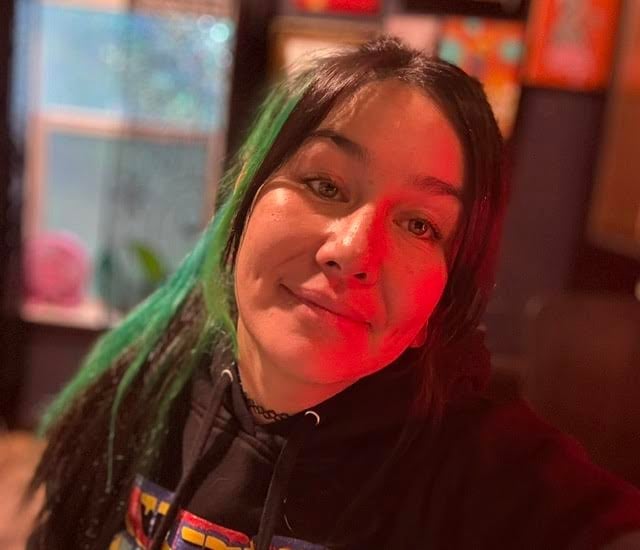 Young person with long dark hair wearing black shirt, illuminated by red and green neon lights in indoor setting