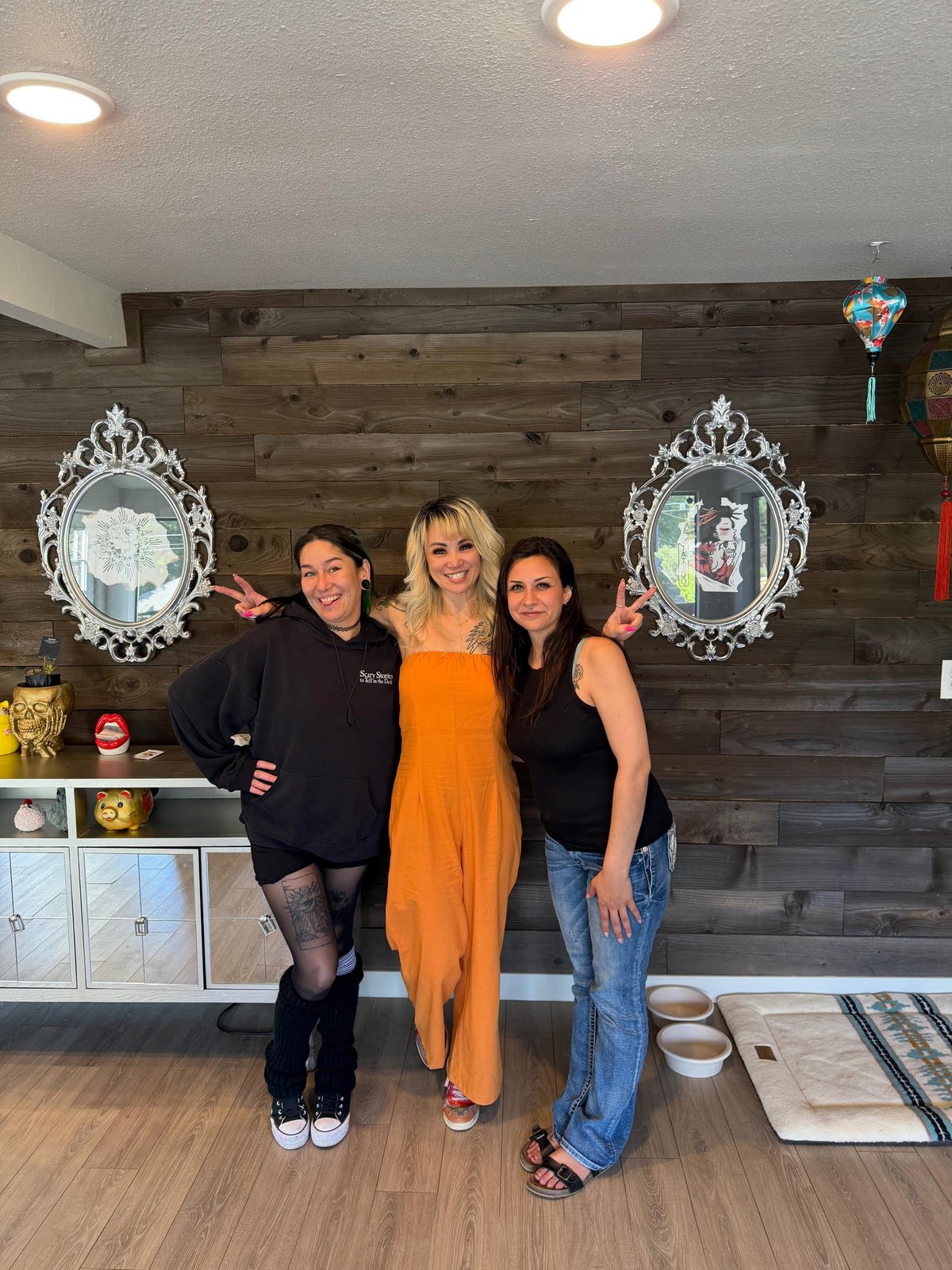 Three women posing together in a modern salon with dark wood accent wall, ornate mirrors, and recessed lighting